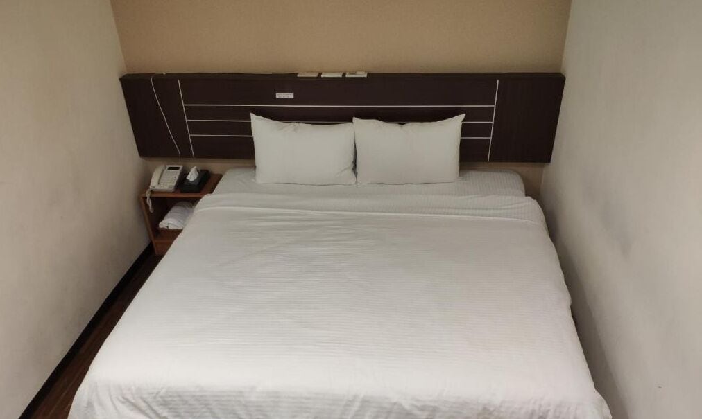 Un letto rifatto con lenzuola bianche, una testiera scura e un comodino con un telefono.