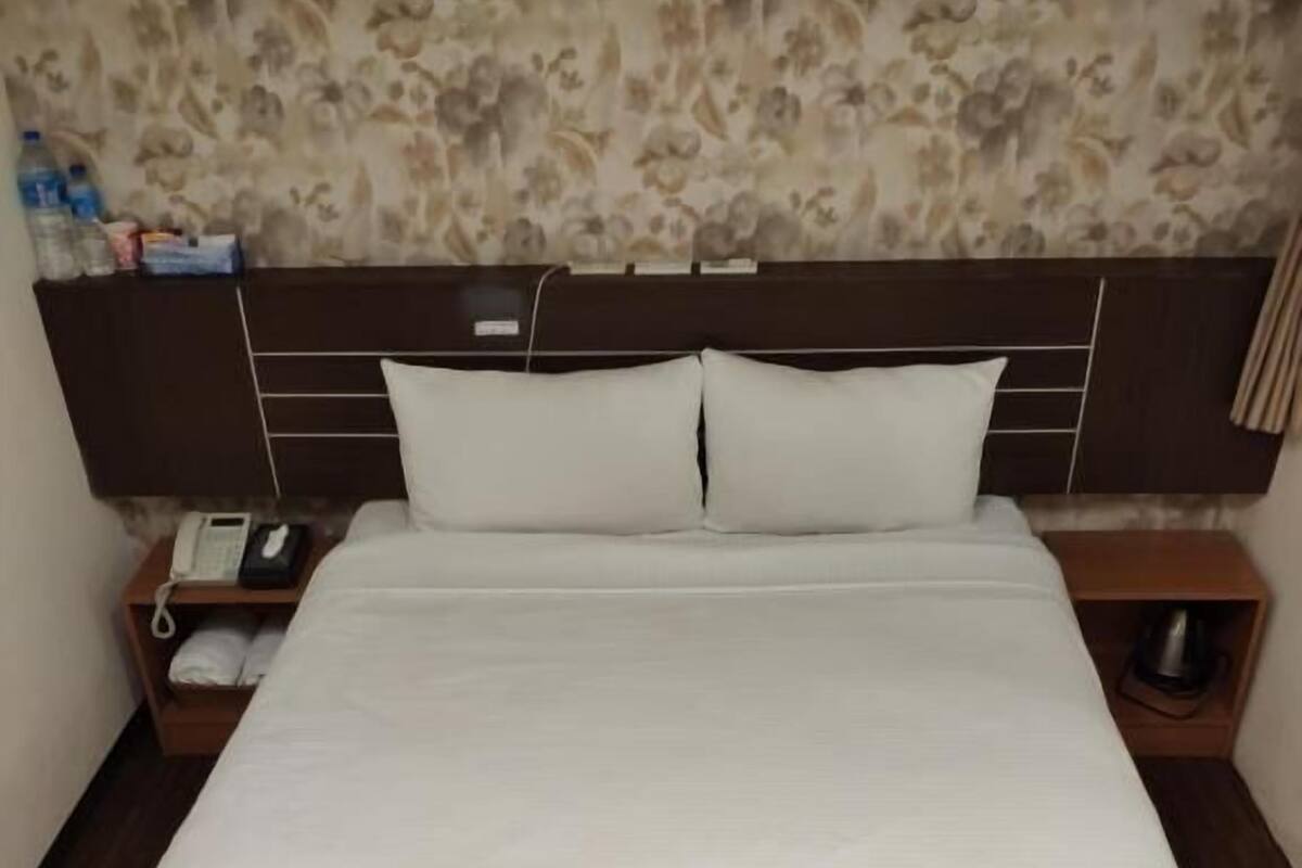 A neatly made bed with white linens, a wooden headboard, and a nightstand with a phone and a bottle.