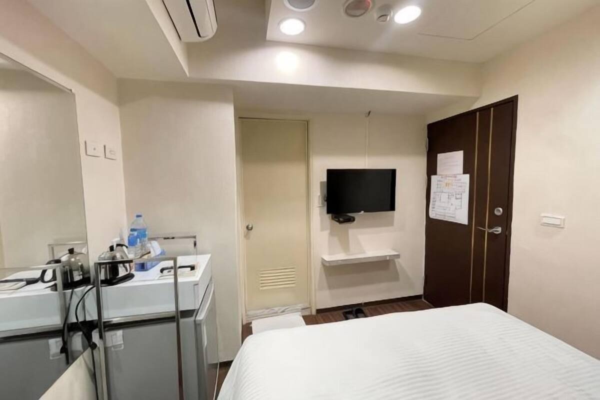 A hotel room with a bed, a desk with a kettle, a flat-screen TV, and a door with a notice.