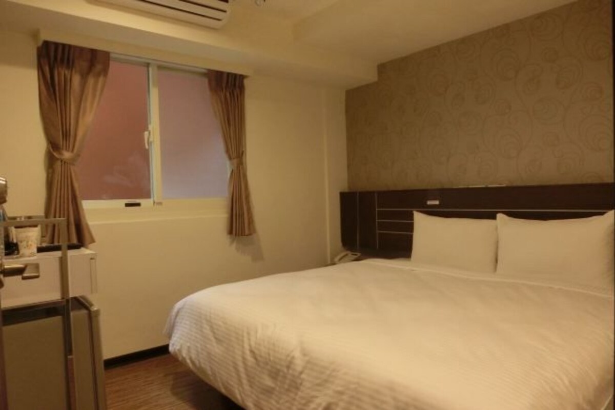 A hotel room with a bed, a desk, a window with curtains, and a headboard.