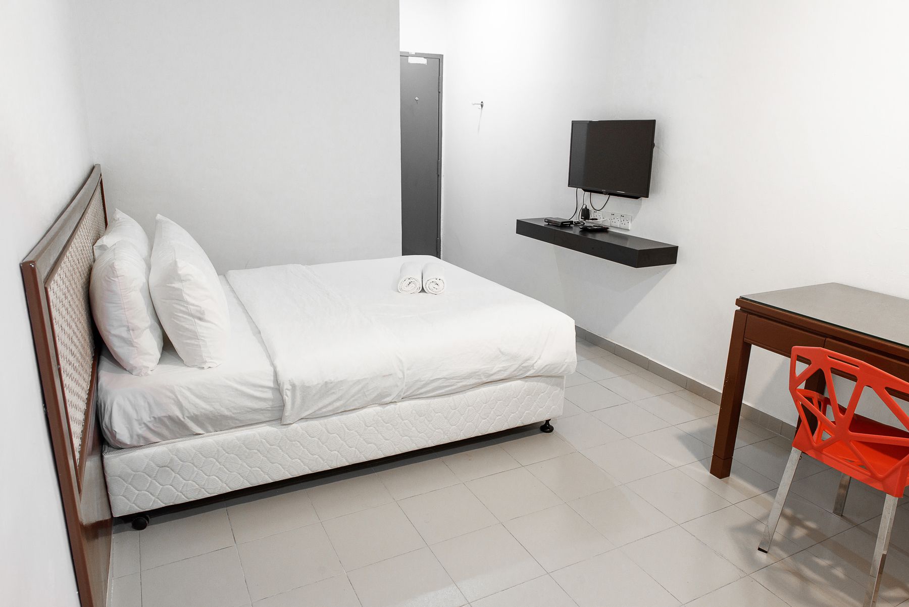 Une chambre avec un lit simple, une tête de lit en bois, un téléviseur à écran plat fixé sur une étagère noire, et une petite table en bois avec une chaise rouge.