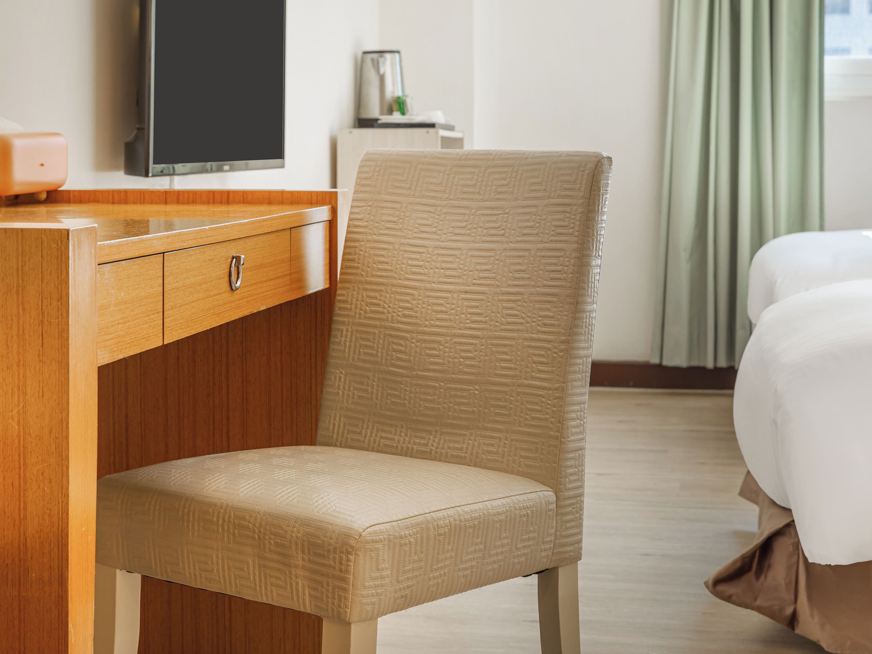 A hotel room with a wooden desk, a beige upholstered chair, a bed with white bedding, and a television on the wall.