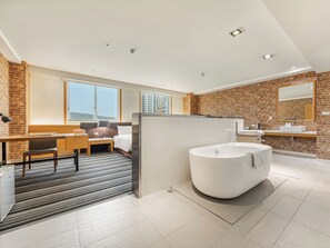 Una habitación de hotel moderna con un ventanal grande, una pared de ladrillo decorativa, una bañera amplia, un escritorio con una silla y una pequeña mesa con una lámpara.