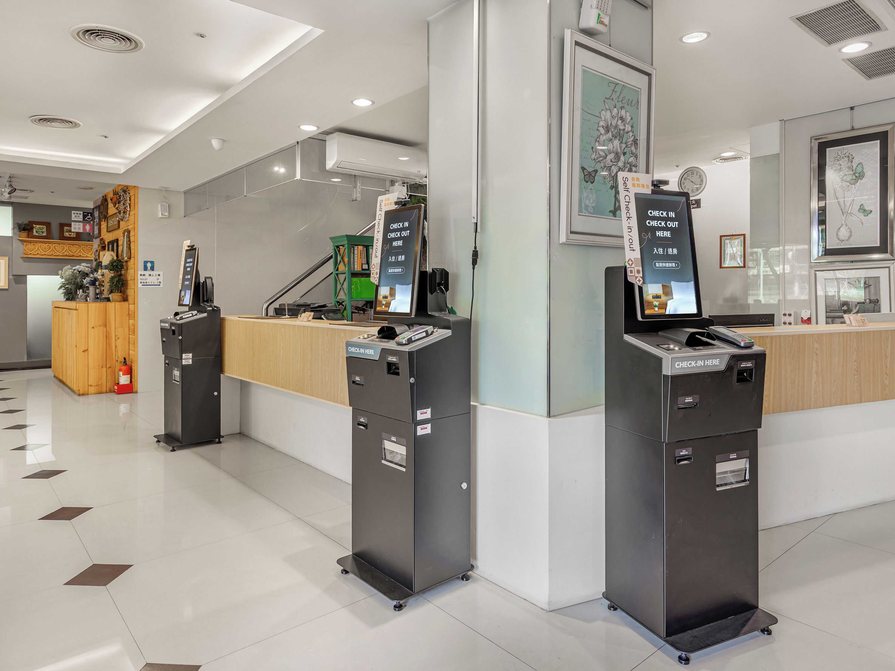 Zona de recepción de hotel con quioscos de registro, mostrador de recepción y una planta verde.