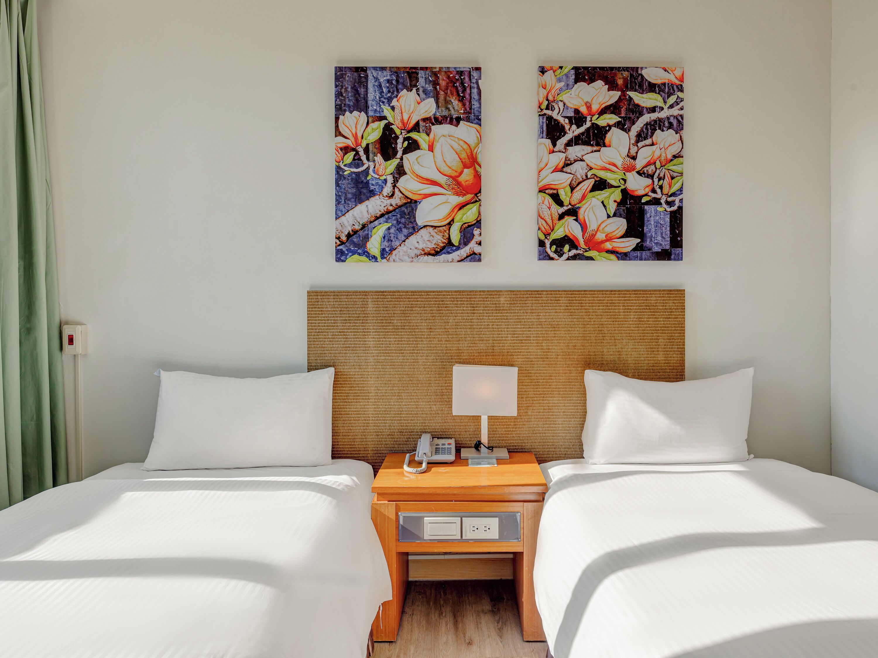 Une chambre d’hôtel avec deux lits, une table de chevet en bois avec une lampe, et deux tableaux aux motifs floraux accrochés au mur.