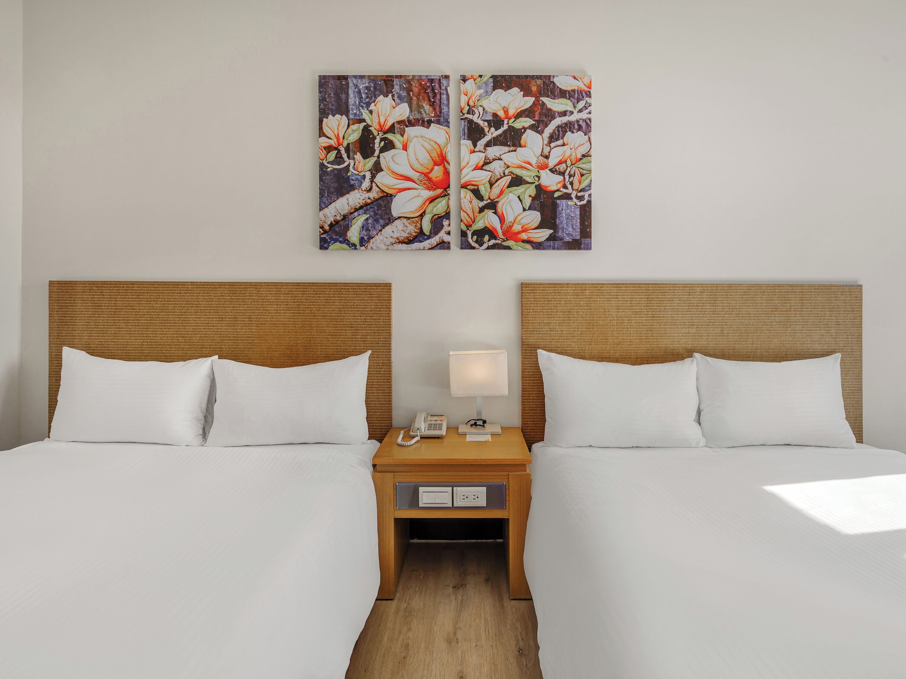 Una habitación de hotel con dos camas, una mesita de noche con una lámpara, y un cuadro de flores en la pared.