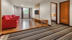Quarto de hotel com piso de madeira, televisão fixada na parede, cama, escrivaninha e área de estar com cadeiras vermelhas.