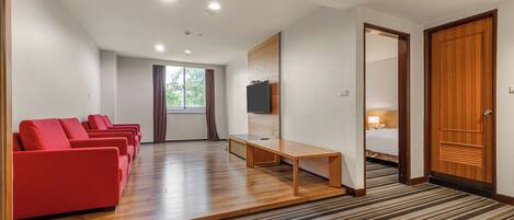 Habitación de hotel con piso de madera, televisor montado en la pared, cama, escritorio y zona de estar con sillas rojas.