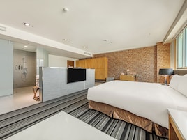 Quarto de hotel moderno com uma cama grande, televisão de tela plana, escrivaninha com luminária, parede de tijolos e área de chuveiro.