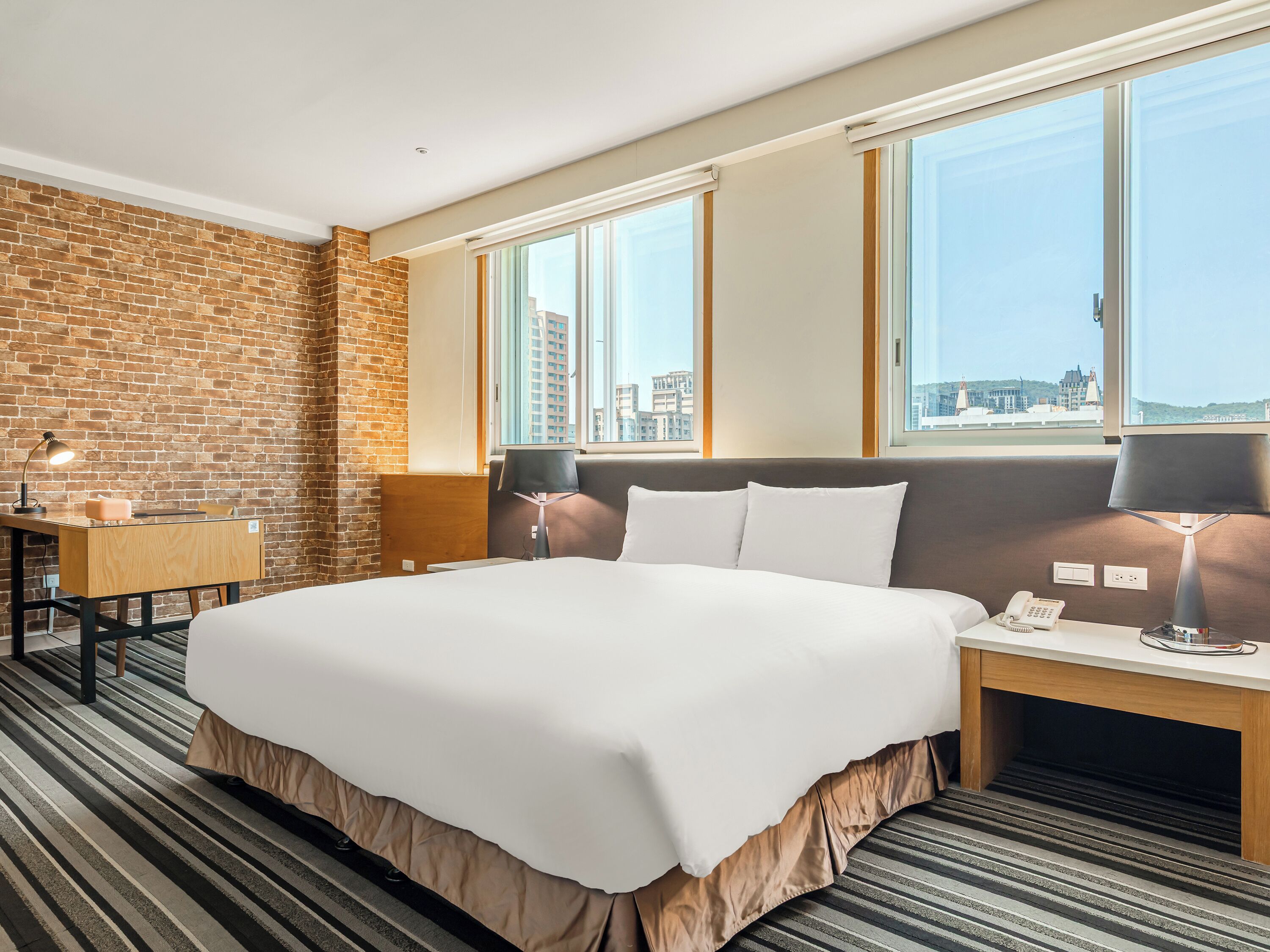A hotel room with a large bed, a desk, a lamp, a phone, and a view of the city through large windows.