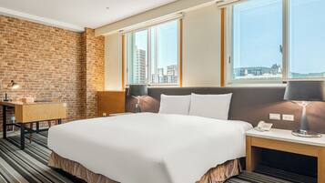 A hotel room with a large bed, a desk, a lamp, a phone, and a view of the city through large windows.
