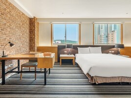 Una camera d'albergo con un muro in mattoni, una scrivania con lampada, un letto con testiera, due comodini con lampade e un'ampia finestra vista città.