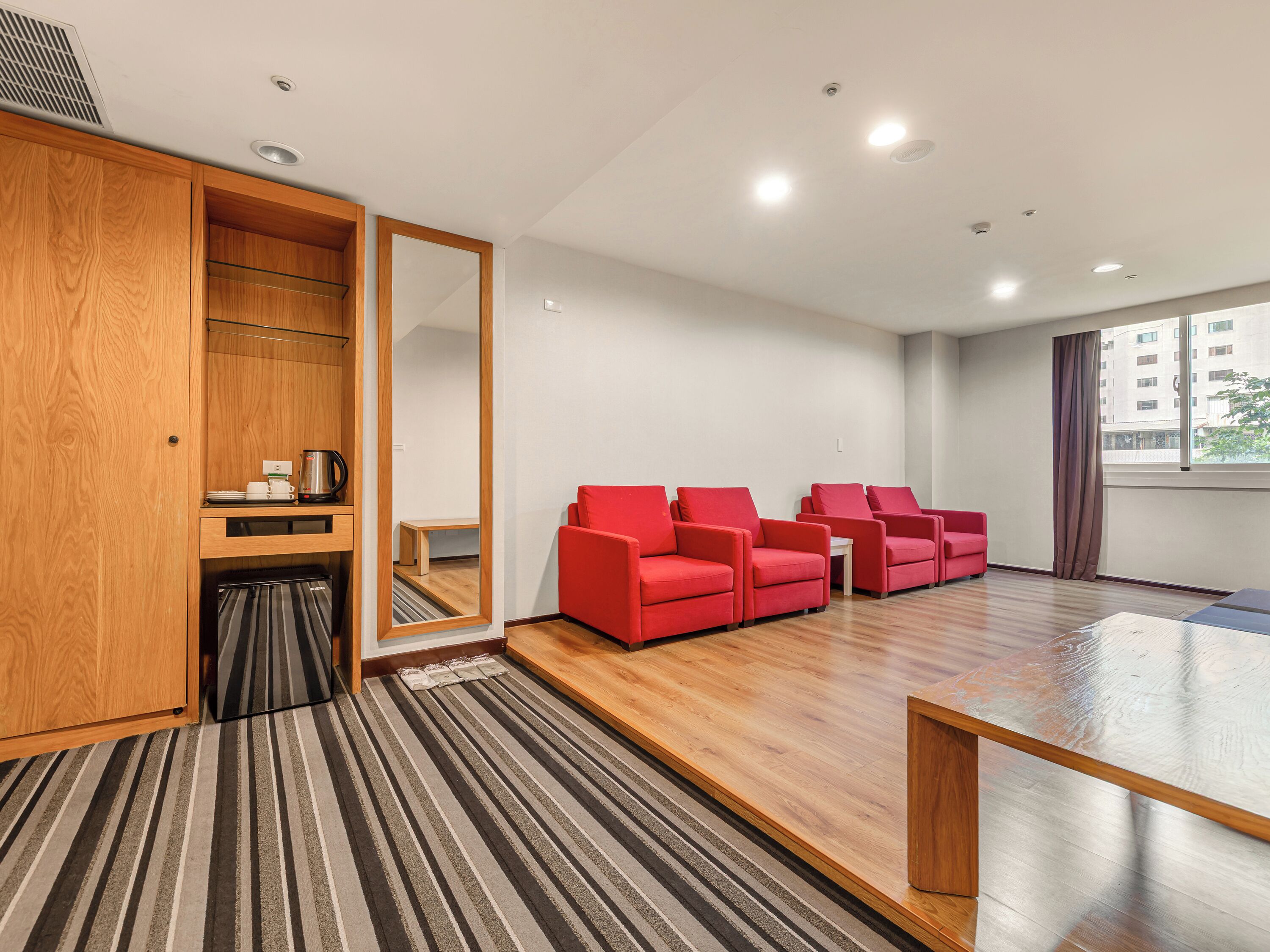 Una habitación de hotel con un armario de madera, alfombra a rayas, sillas rojas, una mesa de centro de madera y un ventanal grande.