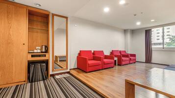 Una habitación de hotel con un armario de madera, alfombra a rayas, sillas rojas, una mesa de centro de madera y un ventanal grande.