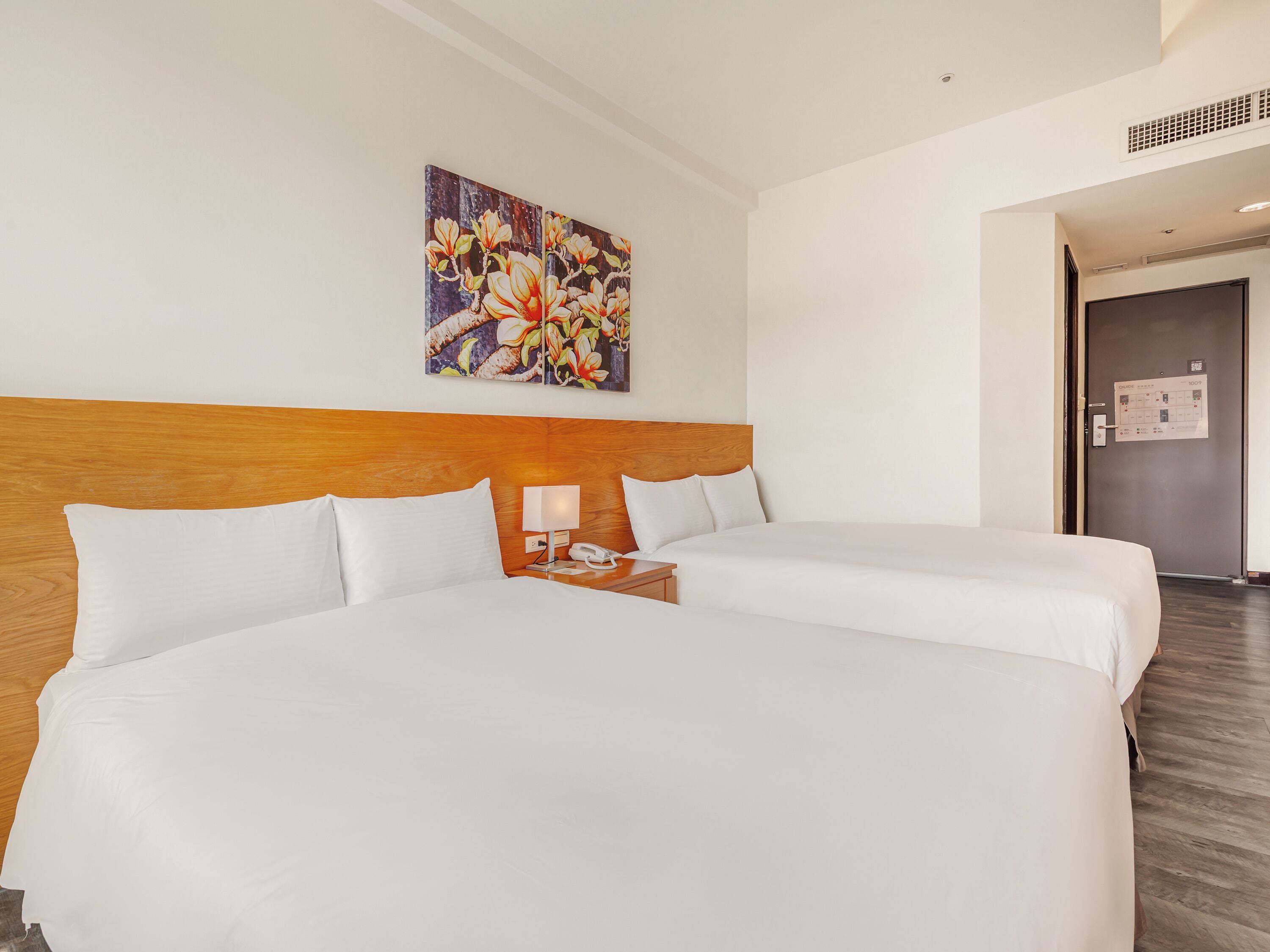 Een hotelkamer met twee bedden, een houten hoofdbord, een nachtkastje met een lamp en een bloemschilderij aan de muur.