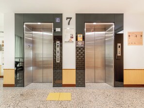 Due porte dell'ascensore con un cartello numero 7, un segnale che indica divieto di fumo e una mappa informativa a parete.