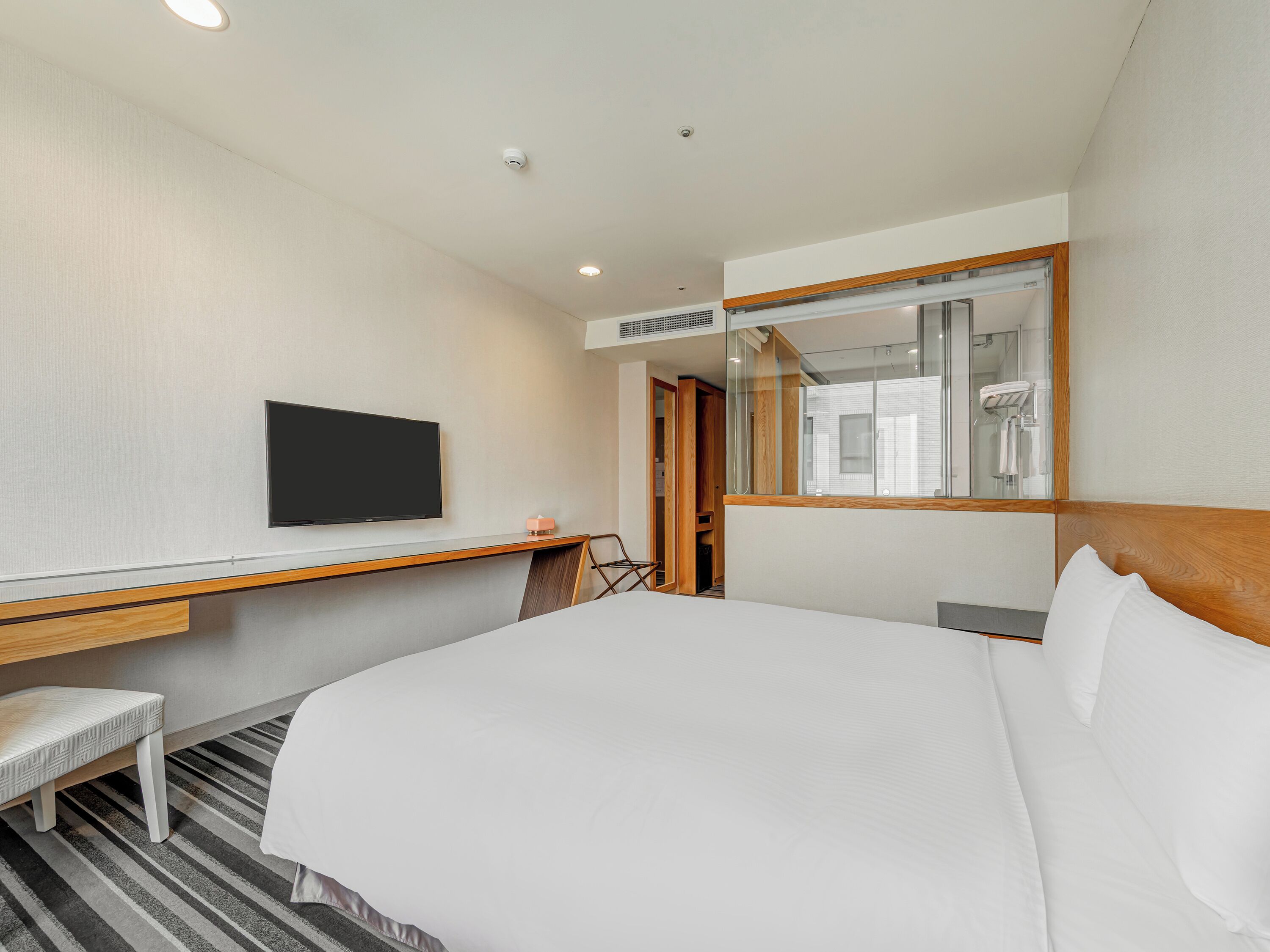 Une chambre d’hôtel équipée d’un lit, d’une télévision, d’un bureau, d’une chaise, d’un miroir et d’une douche que l’on peut voir à travers la porte.