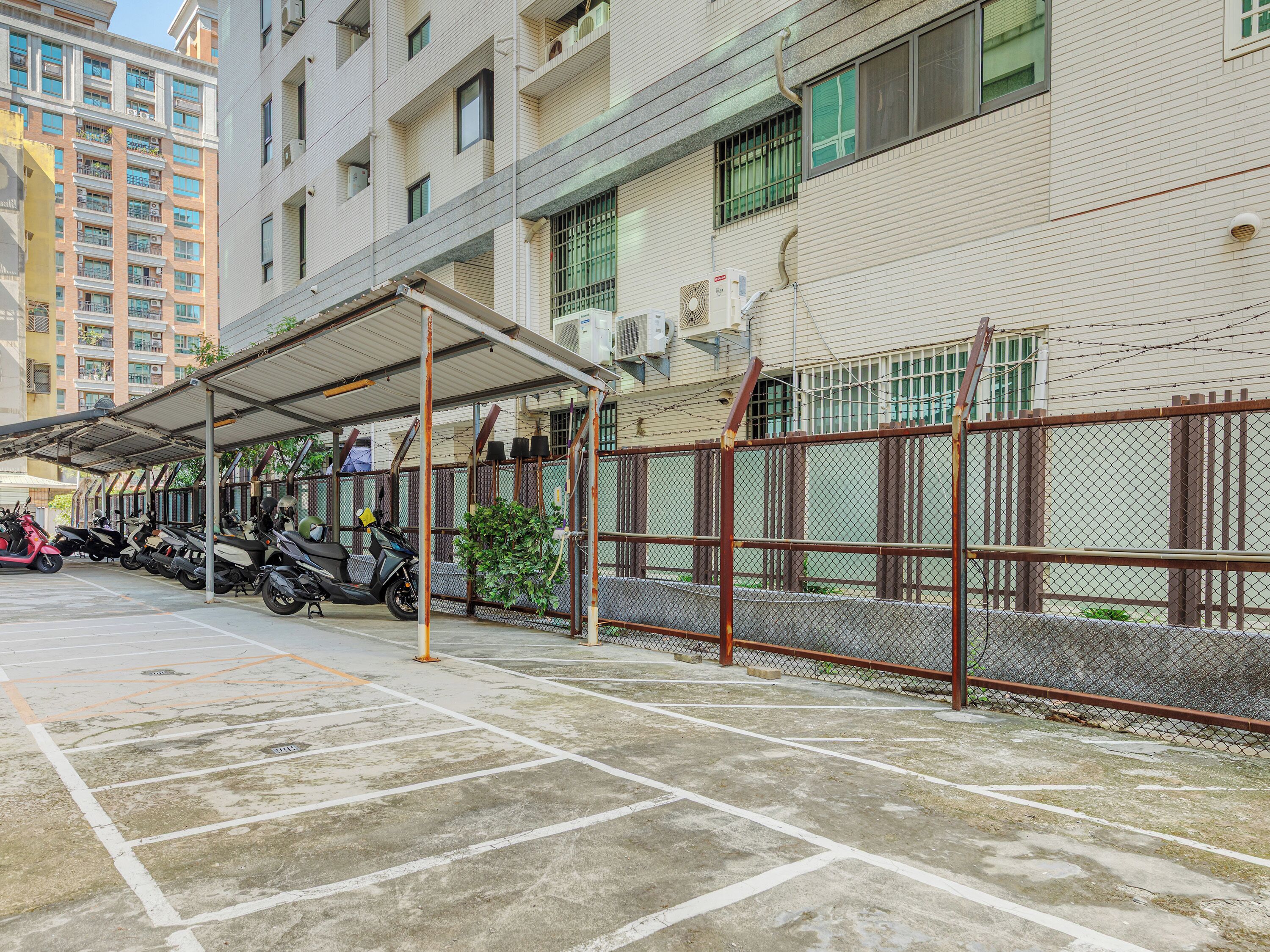 A covered parking area with scooters parked, a chain-link fence, and a building with multiple windows and air conditioning units.