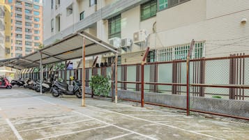 A covered parking area with scooters parked, a chain-link fence, and a building with multiple windows and air conditioning units.