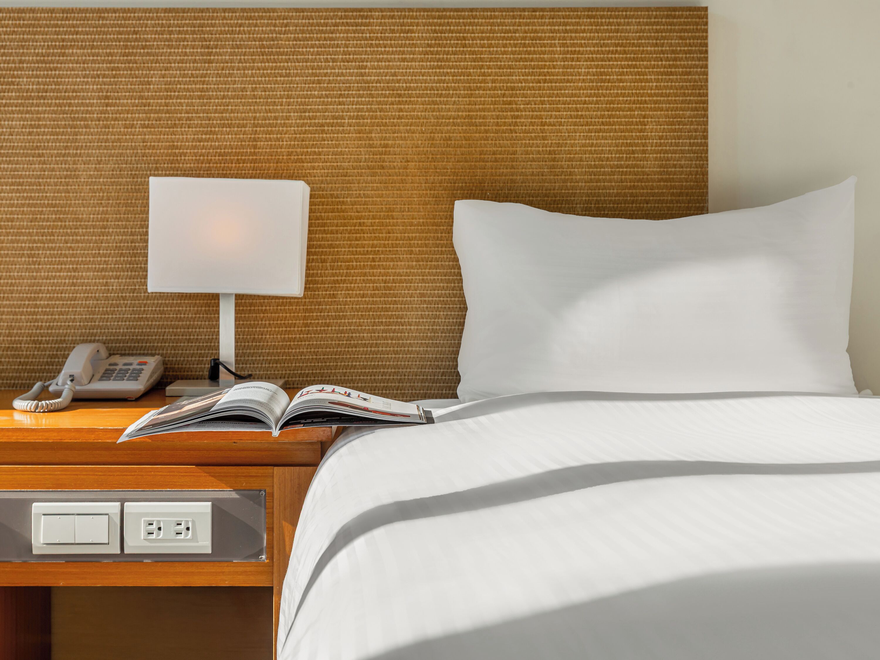 Una habitación de hotel con una cama, una lámpara de noche, un teléfono y un libro en la mesita de noche.