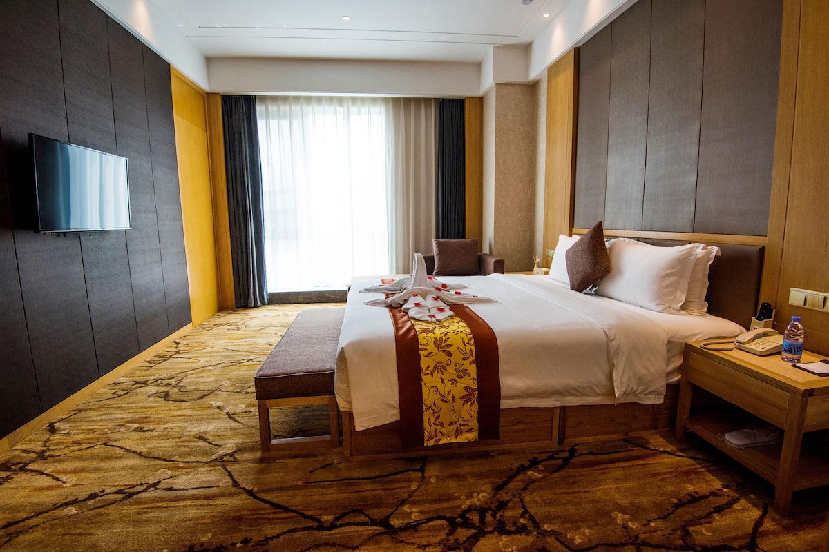 Ein Hotelzimmer mit einem großen Bett, einem Nachttisch mit Telefon und Flasche, einem an der Wand befestigten Flachbildfernseher und einem Fenster mit Vorhängen.