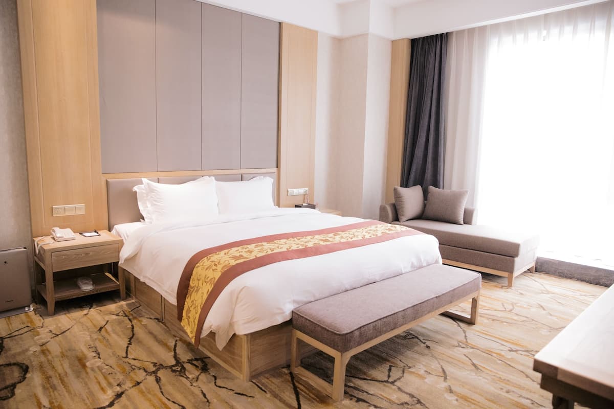 Ein Hotelzimmer mit einem großen Bett, einem Nachttisch mit Telefon, einer Bank, einem Sofa und Blick ins Freie durch ein Fenster.