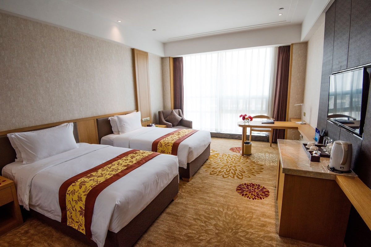 Ein Hotelzimmer mit zwei Betten, einem Schreibtisch, einem Sessel, einem Fernseher und einem großen Fenster mit Vorhängen.