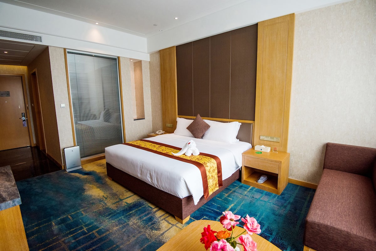 Ein Hotelzimmer mit einem großen Bett, einem Holzkopfende, einem Nachttisch mit Lampe, einem weiteren Holzbeistelltisch, einem braunen Sofa und einem blau gemusterten Teppich.