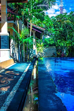 A pool area with a tiled walkway, surrounded by lush greenery and a clear blue sky. - Villa Impian (Canggu)
