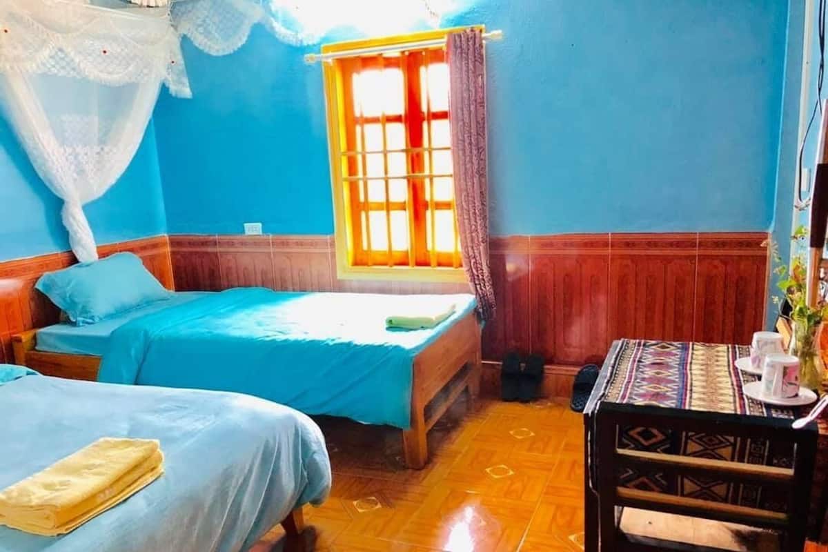 A room with two beds, a window with curtains, a table with cups, and a chair.