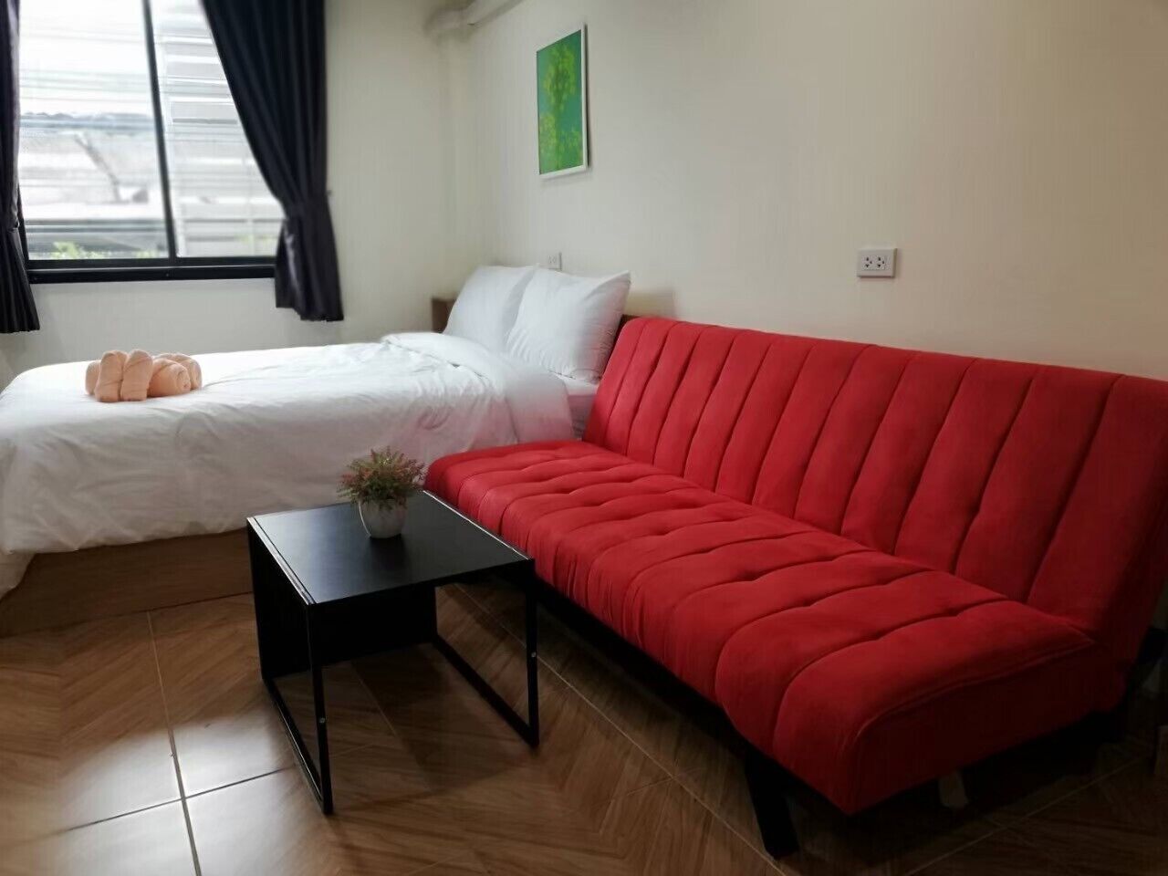A room with a bed, a red sofa, a small black table with a plant, and a window with curtains.