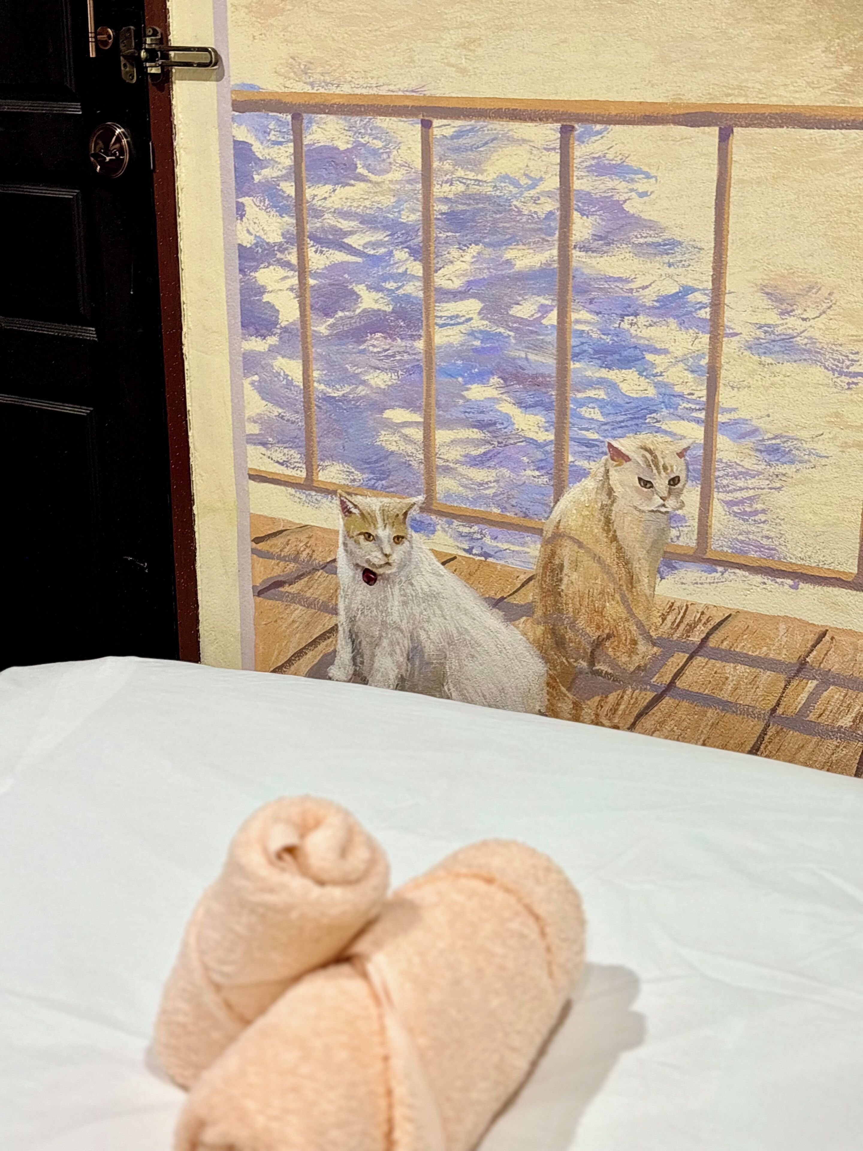 Una cama con toallas enrolladas y dos gatos sentados junto a una ventana con una vista pintada.