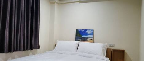 A hotel room with a bed, white bedding, a painting of a beach scene, a window with dark curtains, and a wall-mounted air conditioner.
