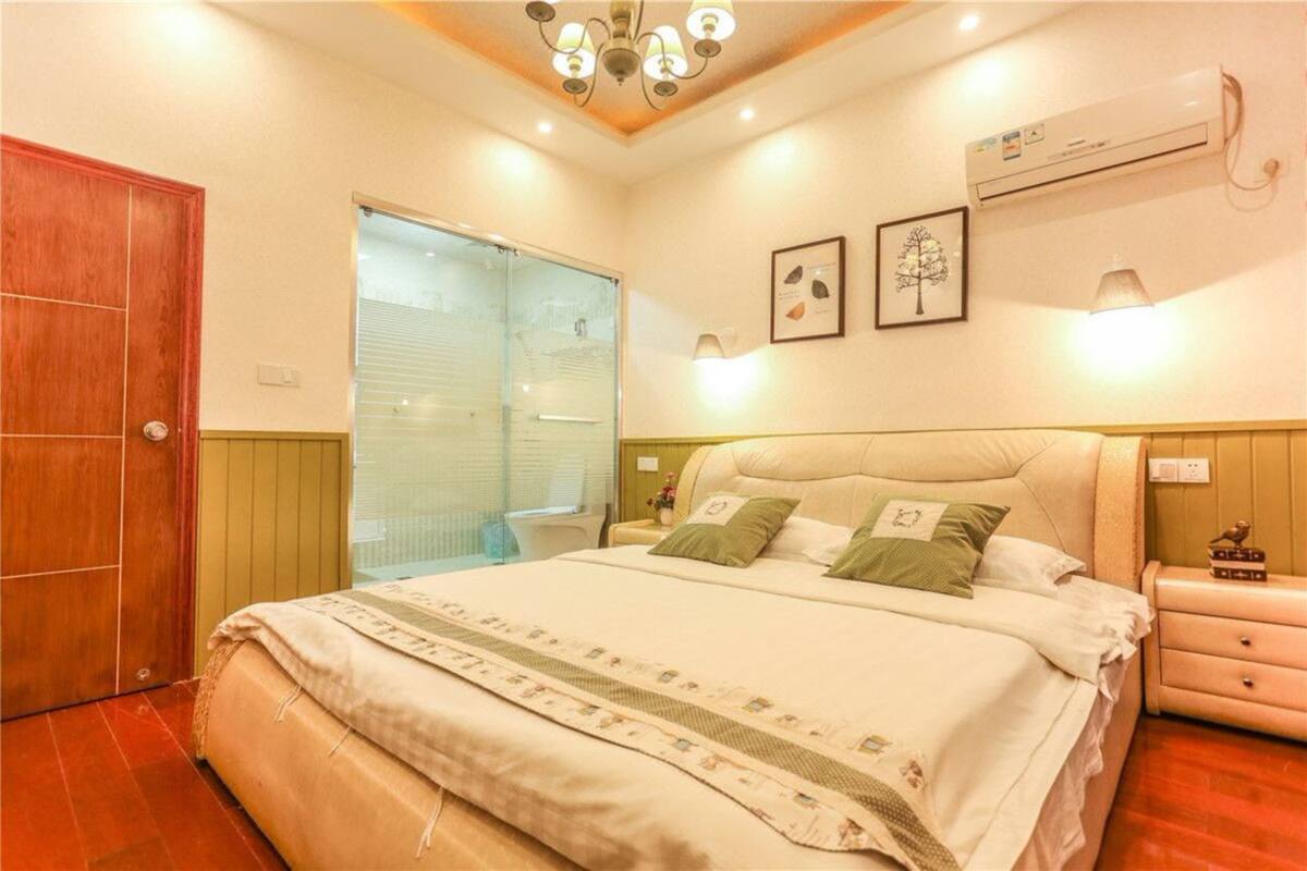 A bedroom with a large bed, a wooden door, a window with blinds, an air conditioning unit, and a nightstand with a lamp.