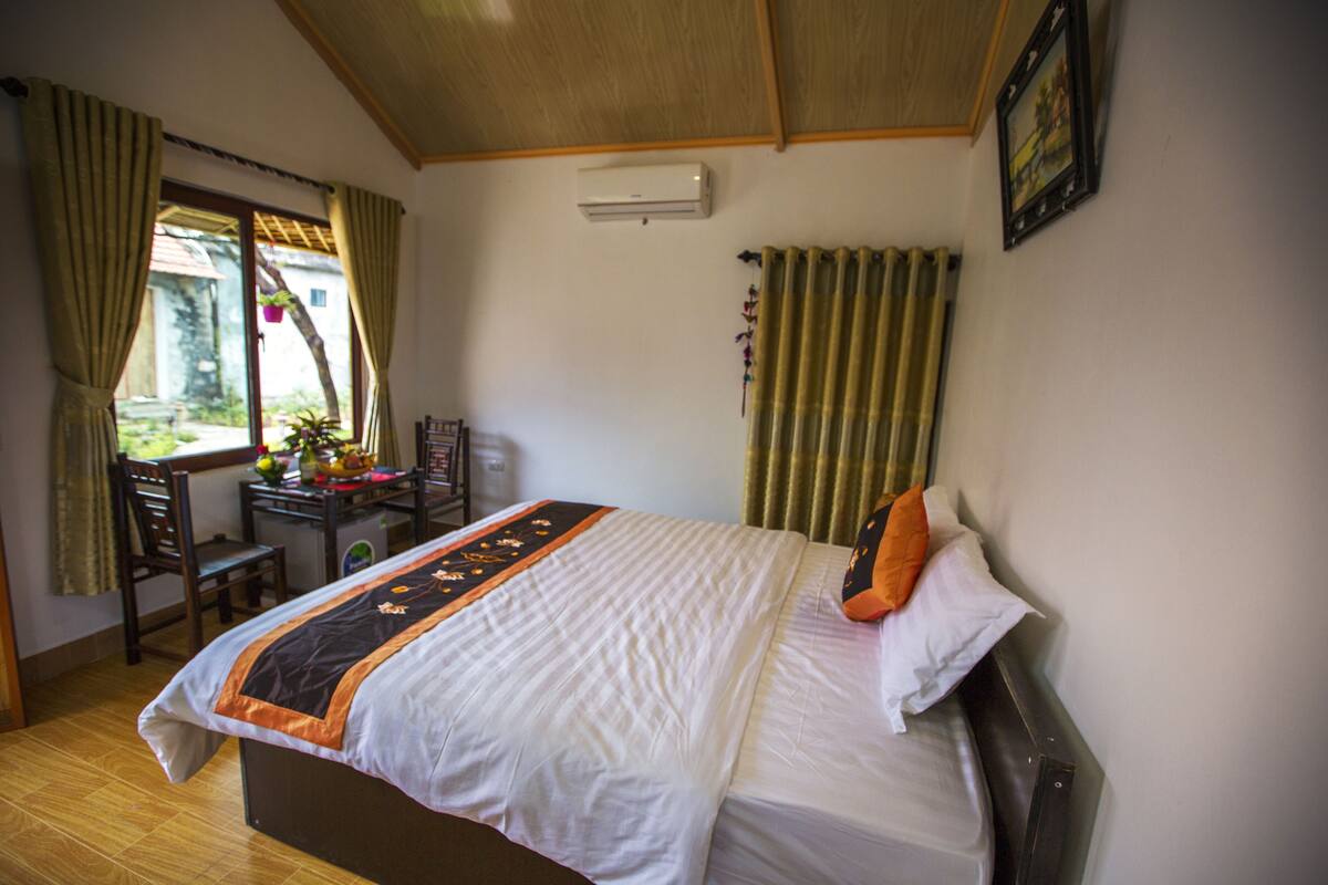 A neatly made bed with a white and black bedspread, a single orange pillow, and a wooden headboard. There is a small table with a chair and a potted plant to the left. An air conditioning unit is mounted on the wall above the bed.