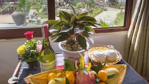 Et bord med frugtkurv, flaske og en potteplante ved et vindue med udsigt til en gårdsplads.