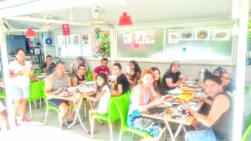 A group of people dining at an outdoor restaurant with green chairs and tables, a menu board, and a ceiling fan.