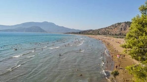 A beach with clear blue water, people swimming and walking along the shore, and mountains in the background. - Karaca Apart Hotel (Ortaca)