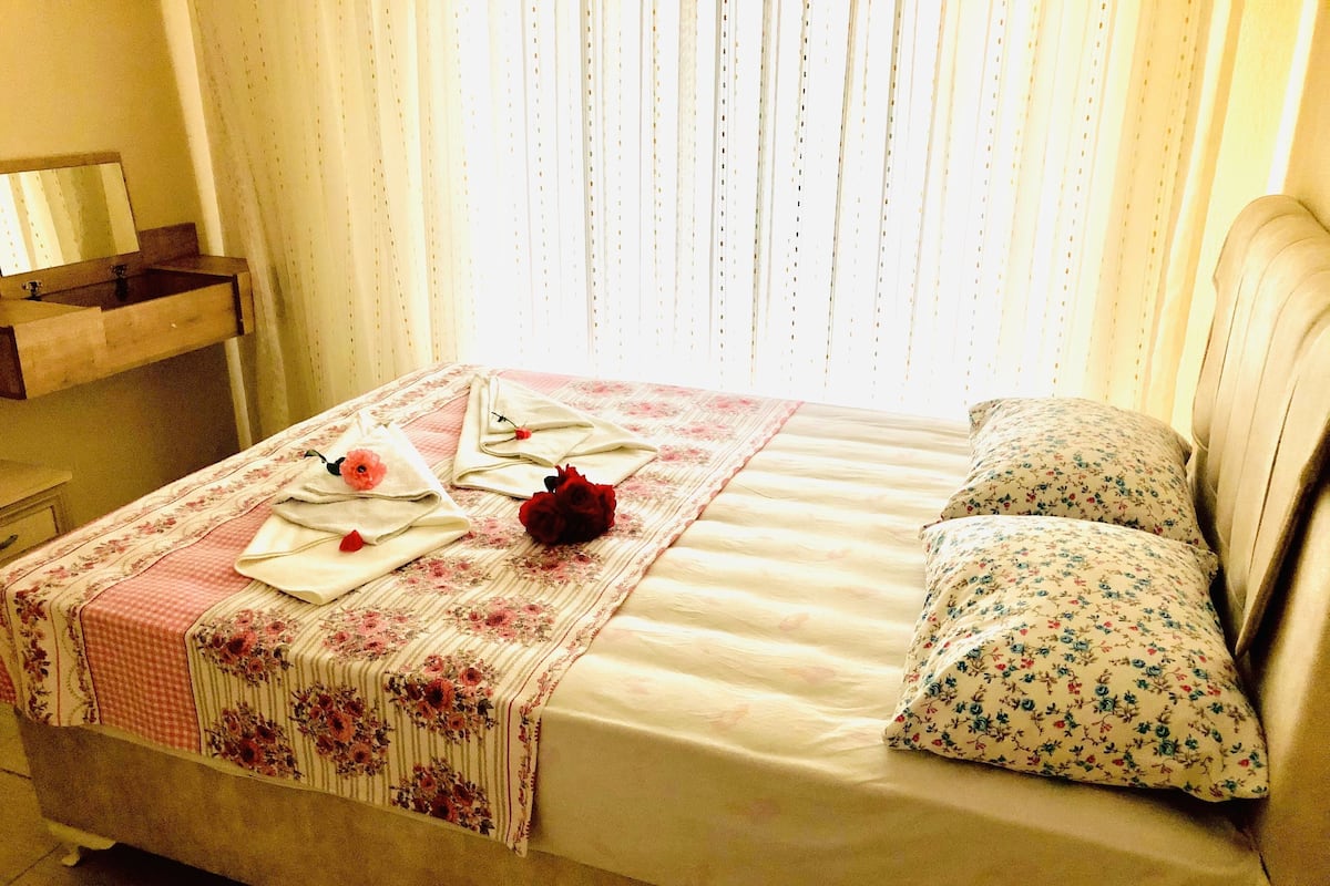 A neatly made bed with floral bedding, a bedside table with a mirror, and a window with vertical blinds.