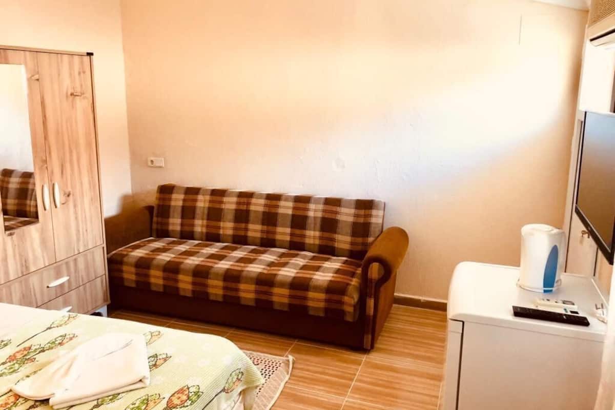 A room with a sofa, a bed with a floral bedspread, a wooden wardrobe, a small white cabinet with a television and a kettle.