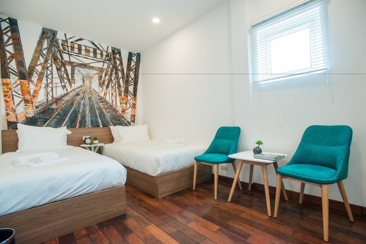 Quarto de hotel com uma cama, duas cadeiras, uma mesinha e uma obra de arte na parede.
