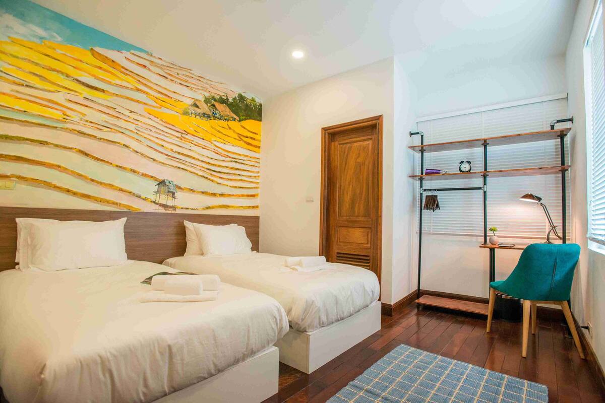 Quarto de hotel com duas camas, uma porta de madeira, uma prateleira com objetos e uma cadeira.