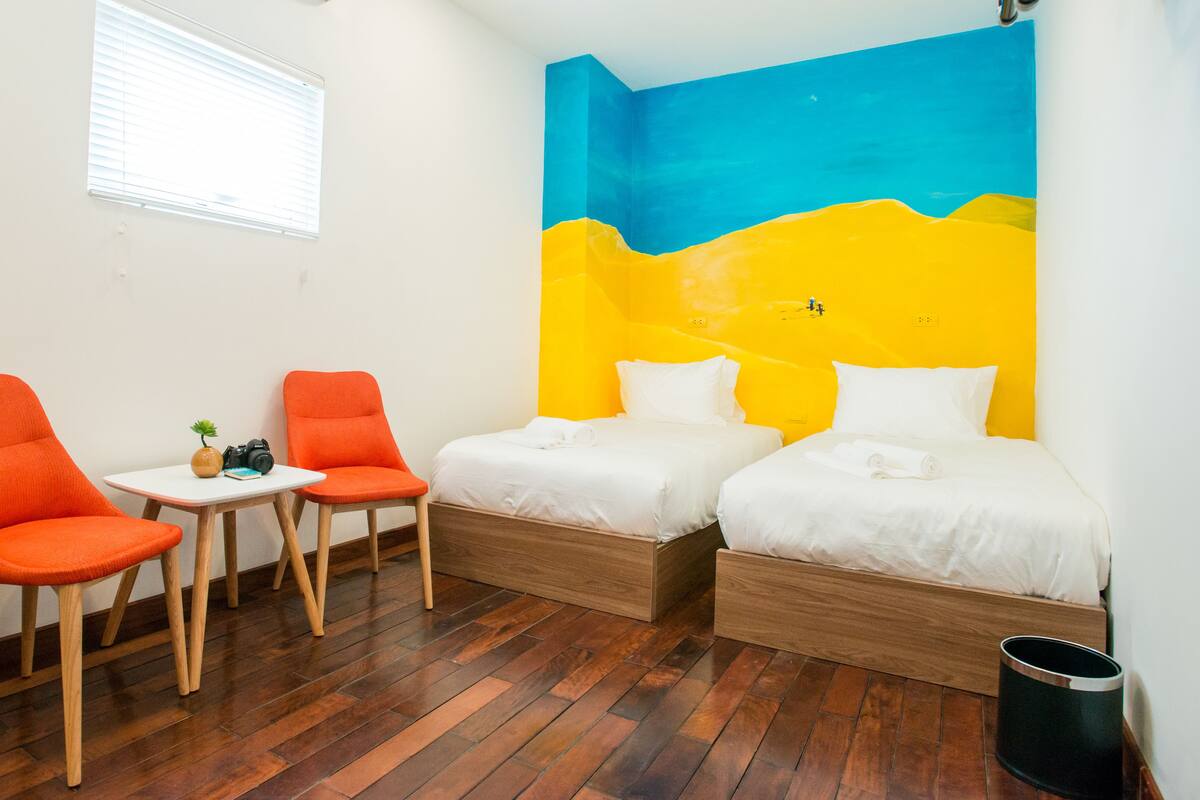 Quarto com duas camas, duas cadeiras laranja, uma mesinha e uma janela com persianas.