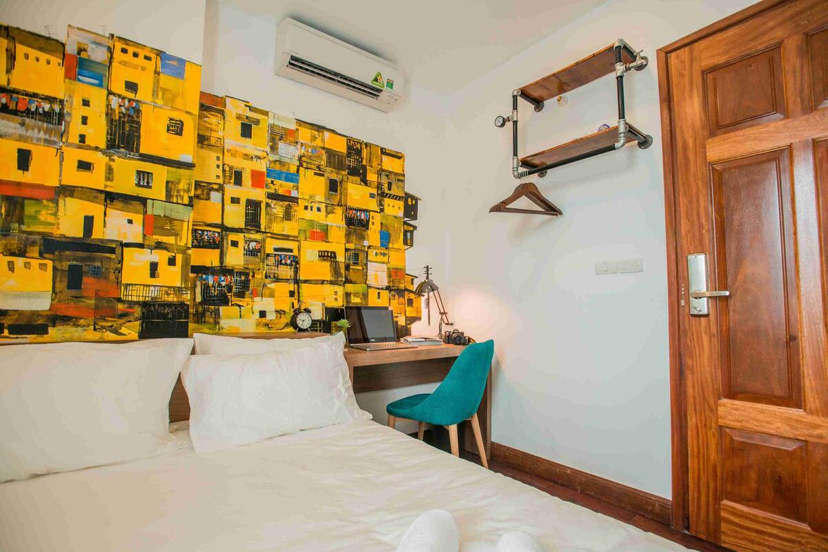 Quarto de hotel com uma cama, uma escrivaninha com cadeira, uma porta de madeira e uma parede decorada com caixas empilhadas.