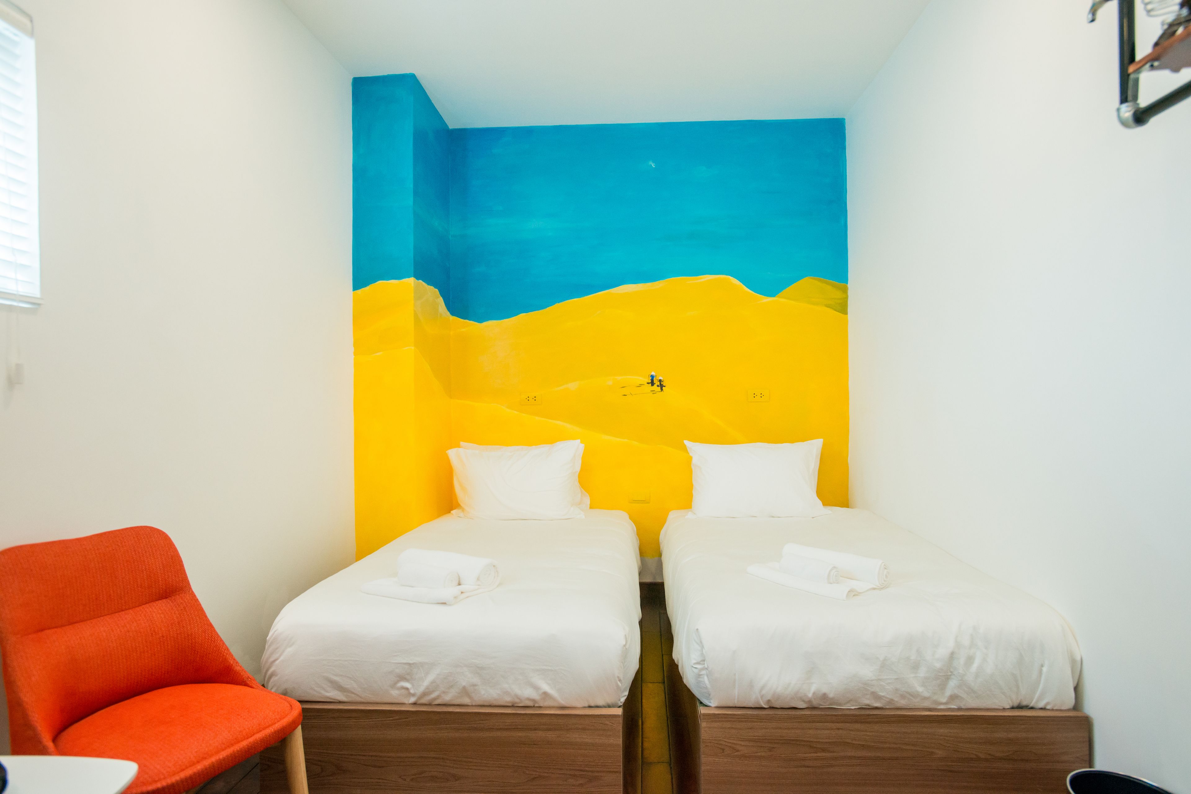Une chambre avec deux lits, un fauteuil orange et un mur orné d’une fresque bleue et jaune.