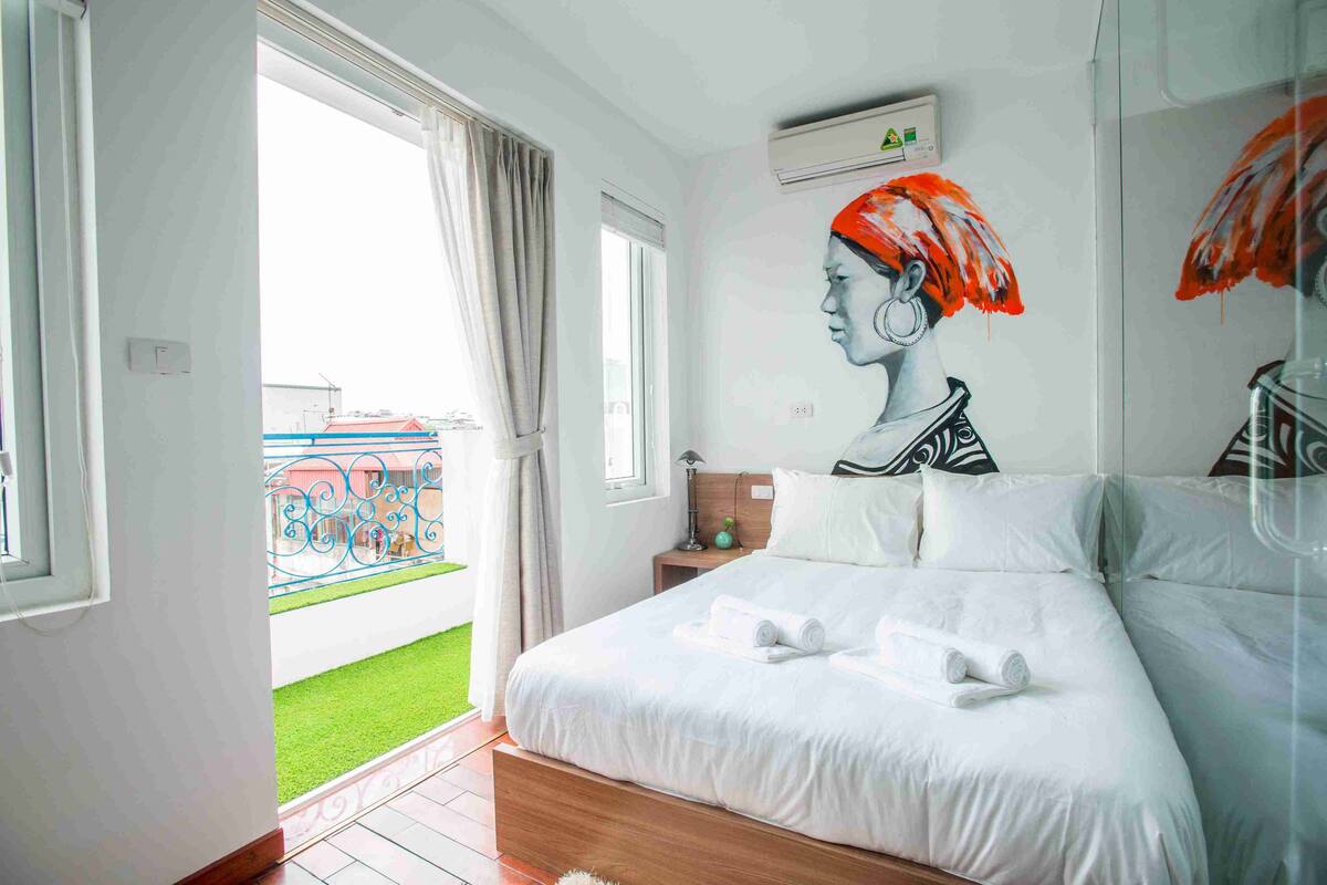 Quarto com cama grande, mesa de cabeceira, ar condicionado fixado na parede e vista para uma varanda com guarda-corpo.