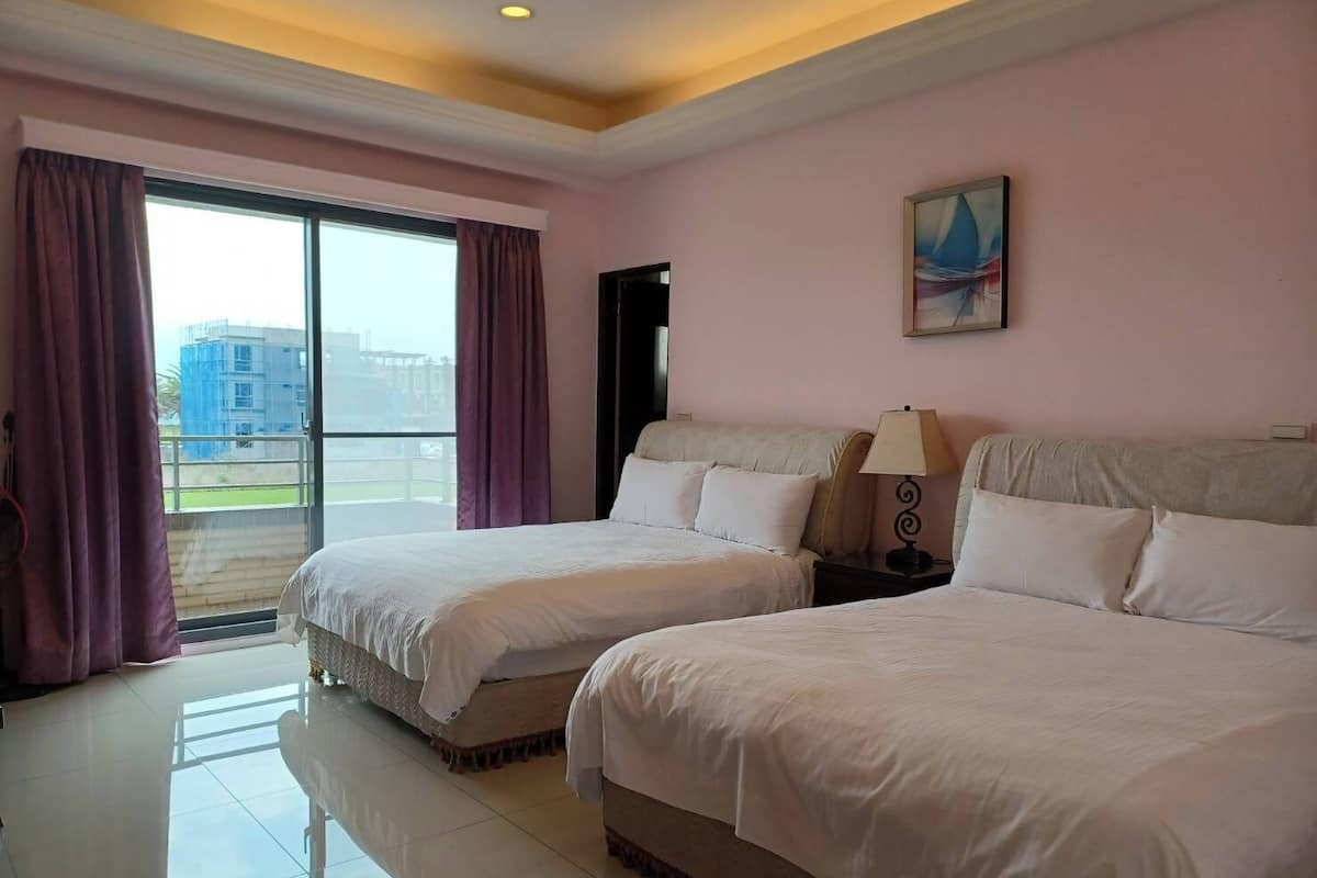 A hotel room with two beds, a balcony with curtains, a painting on the wall, and a view of buildings outside.