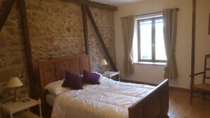 A bedroom with a stone wall, a wooden bed, a nightstand, a lamp, a window with curtains, and a chair. - Gîtes de Campagne (Fursac)