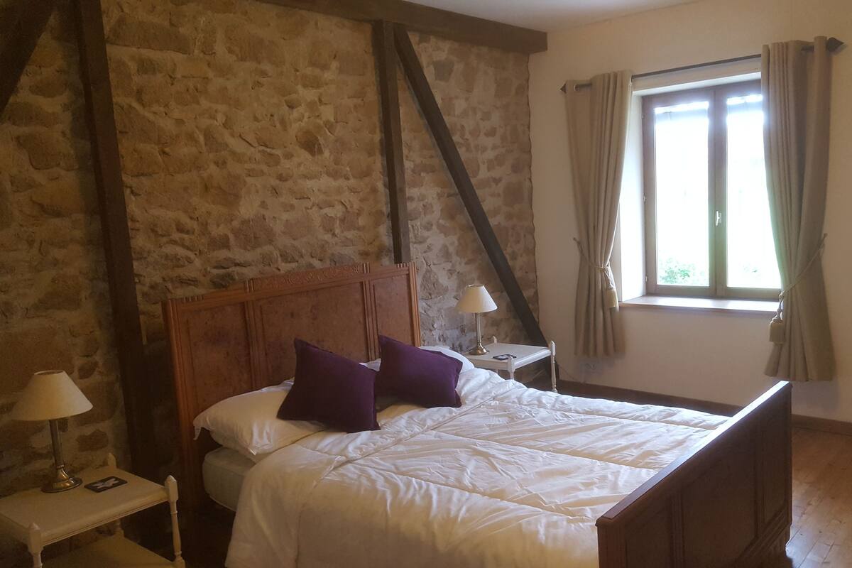 A bedroom with a stone wall, a wooden bed, a nightstand, a lamp, a window with curtains, and a chair.