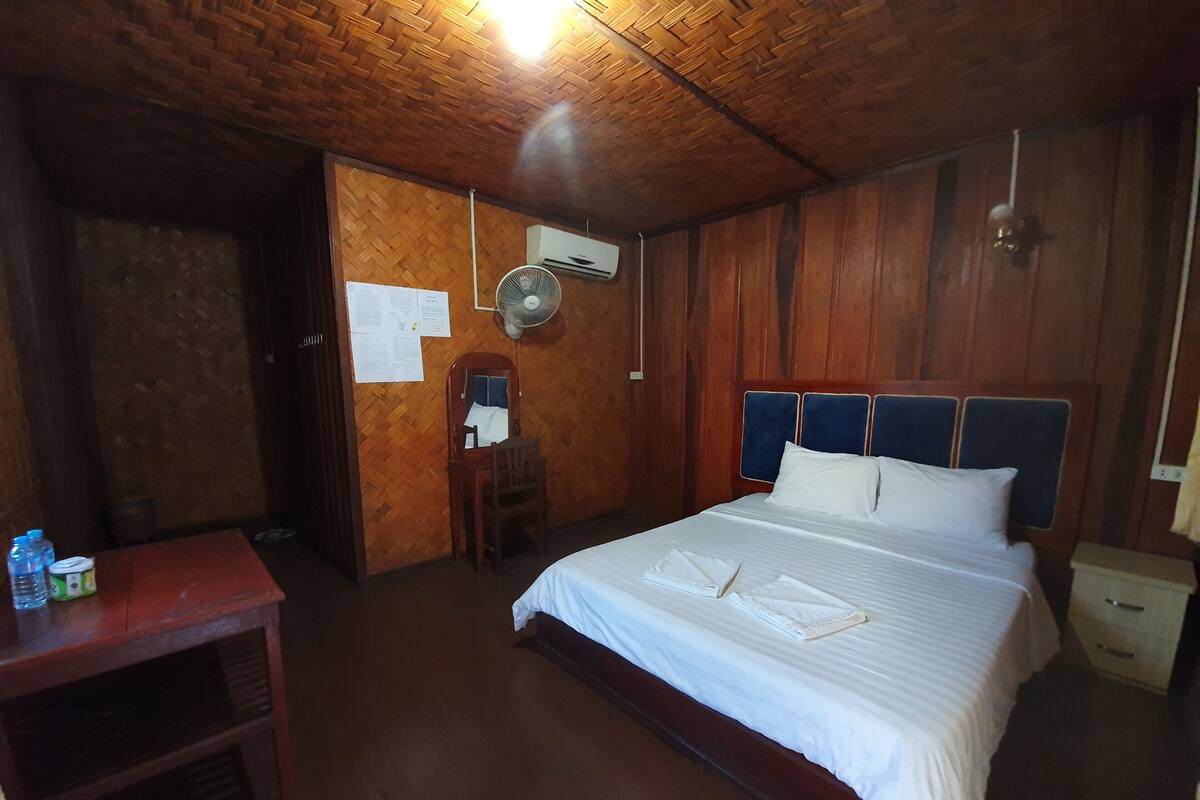 Een kamer met houten lambrisering, een bed, een bureau, een stoel en een ventilator.
